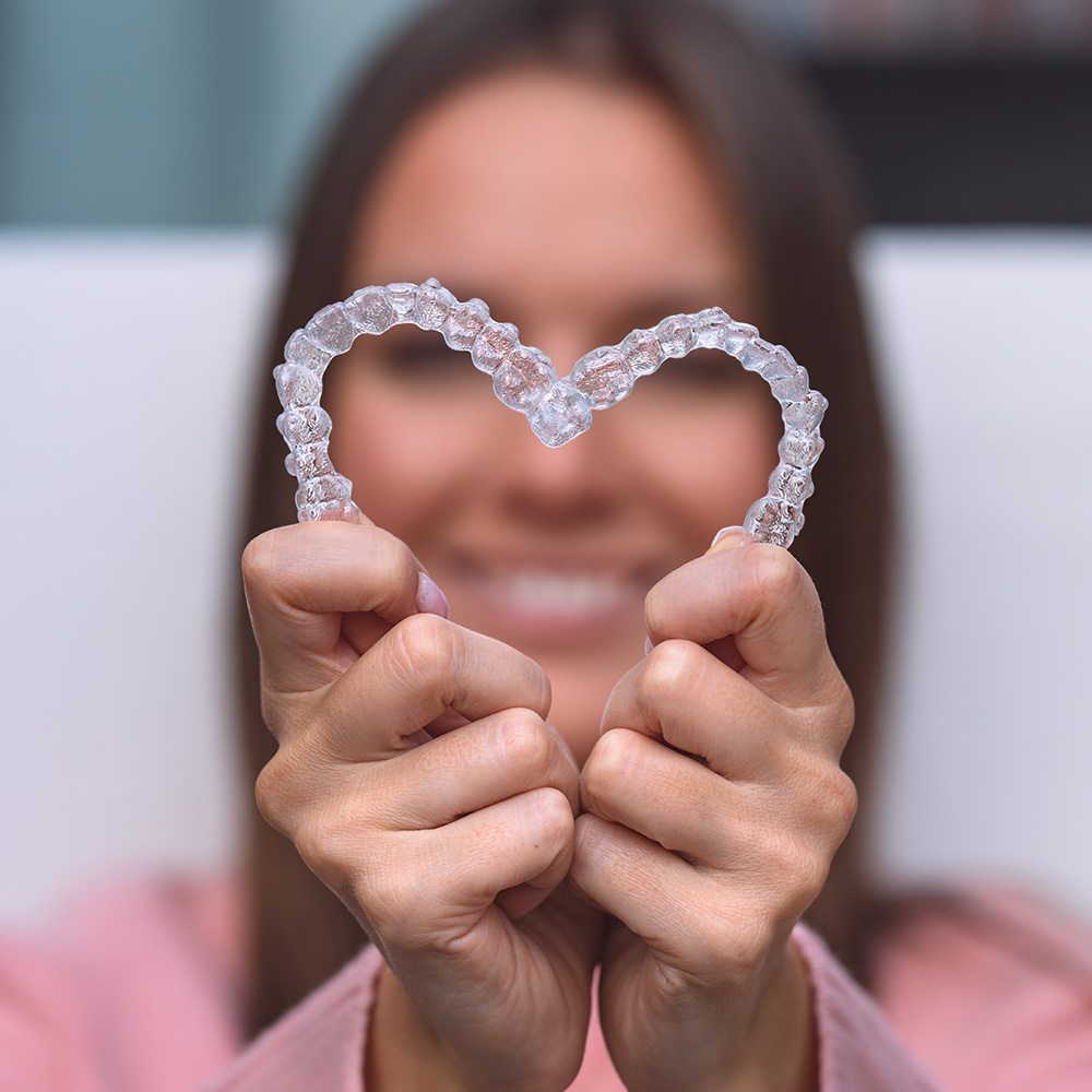 The image shows a person holding up a pair of clear braces with their hands forming a heart shape, against a blurred background.